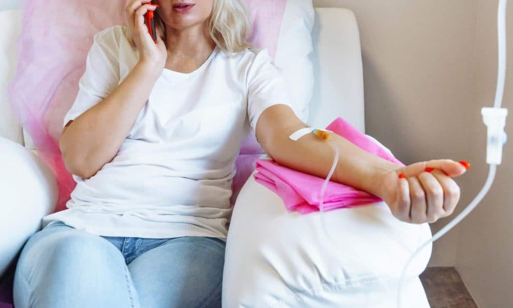 Woman receiving intravenous therapy while on a phone call, showcasing the IV drip and a comfortable setting with pink drapes.